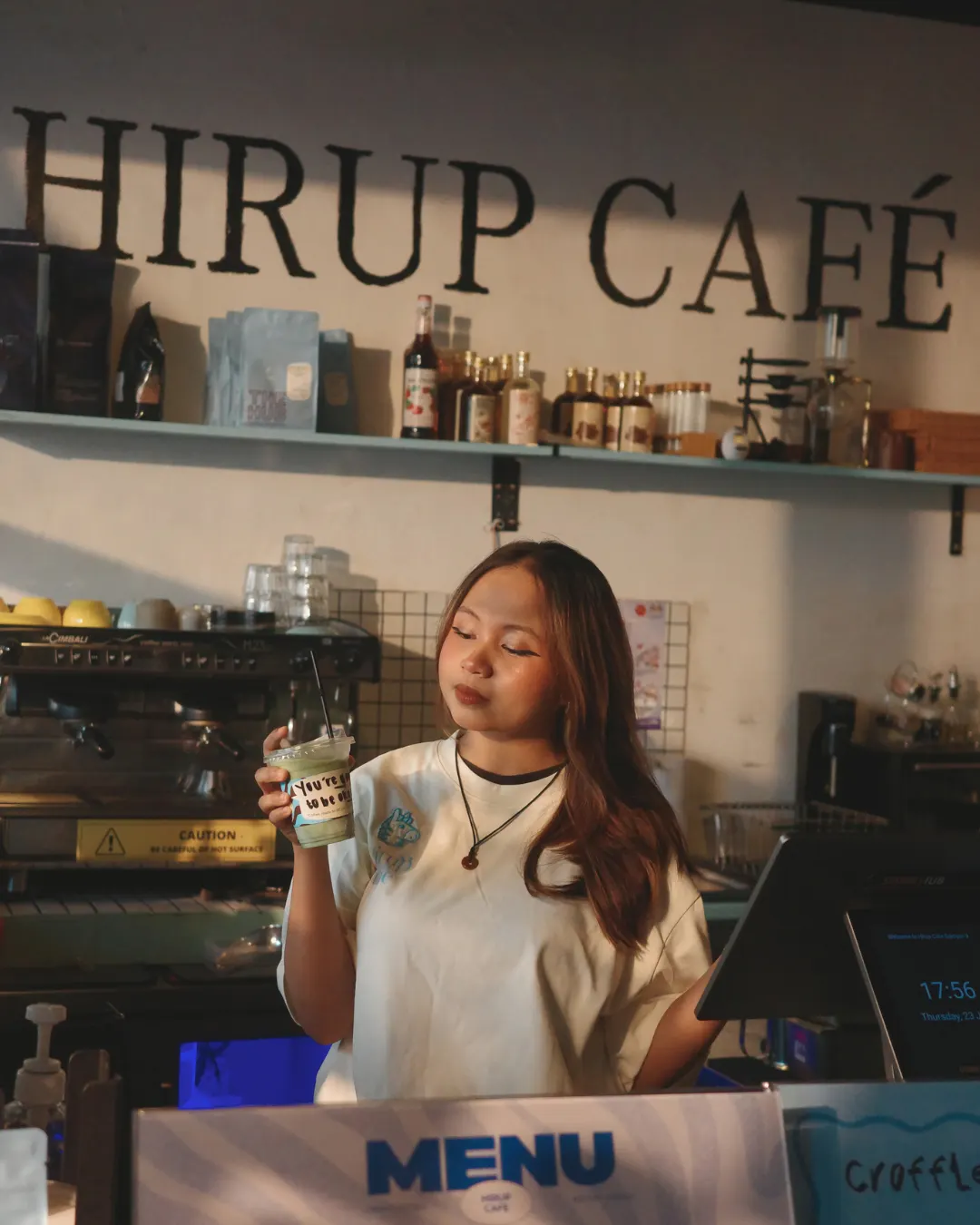 A Hirup barista holding a matcha under the HIRUP CAFÉ sign
